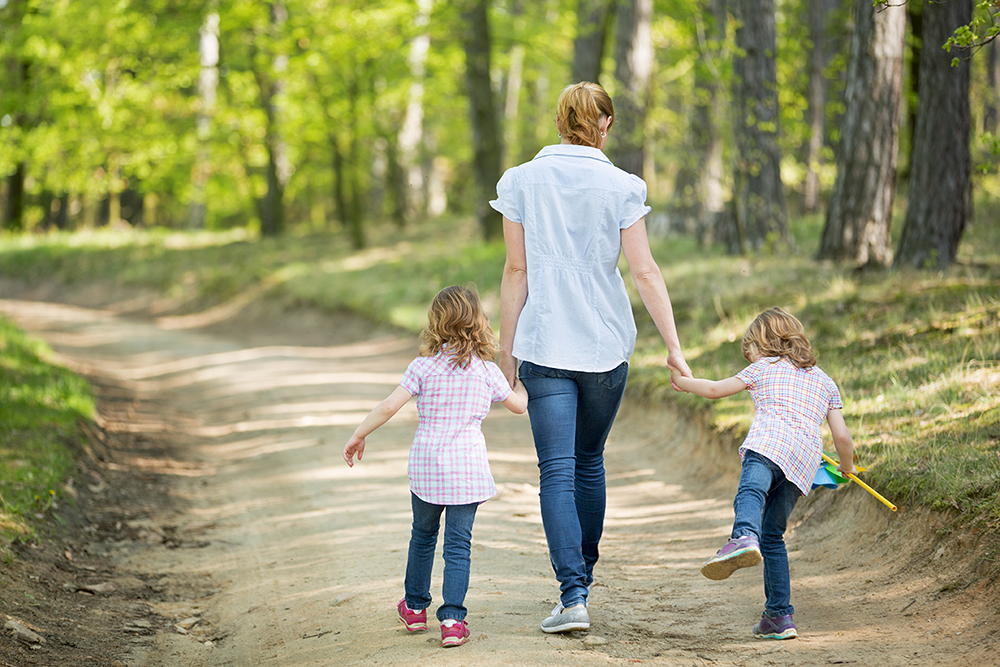 An image of a mother holding hands with her two children walking on a path through a forest. 