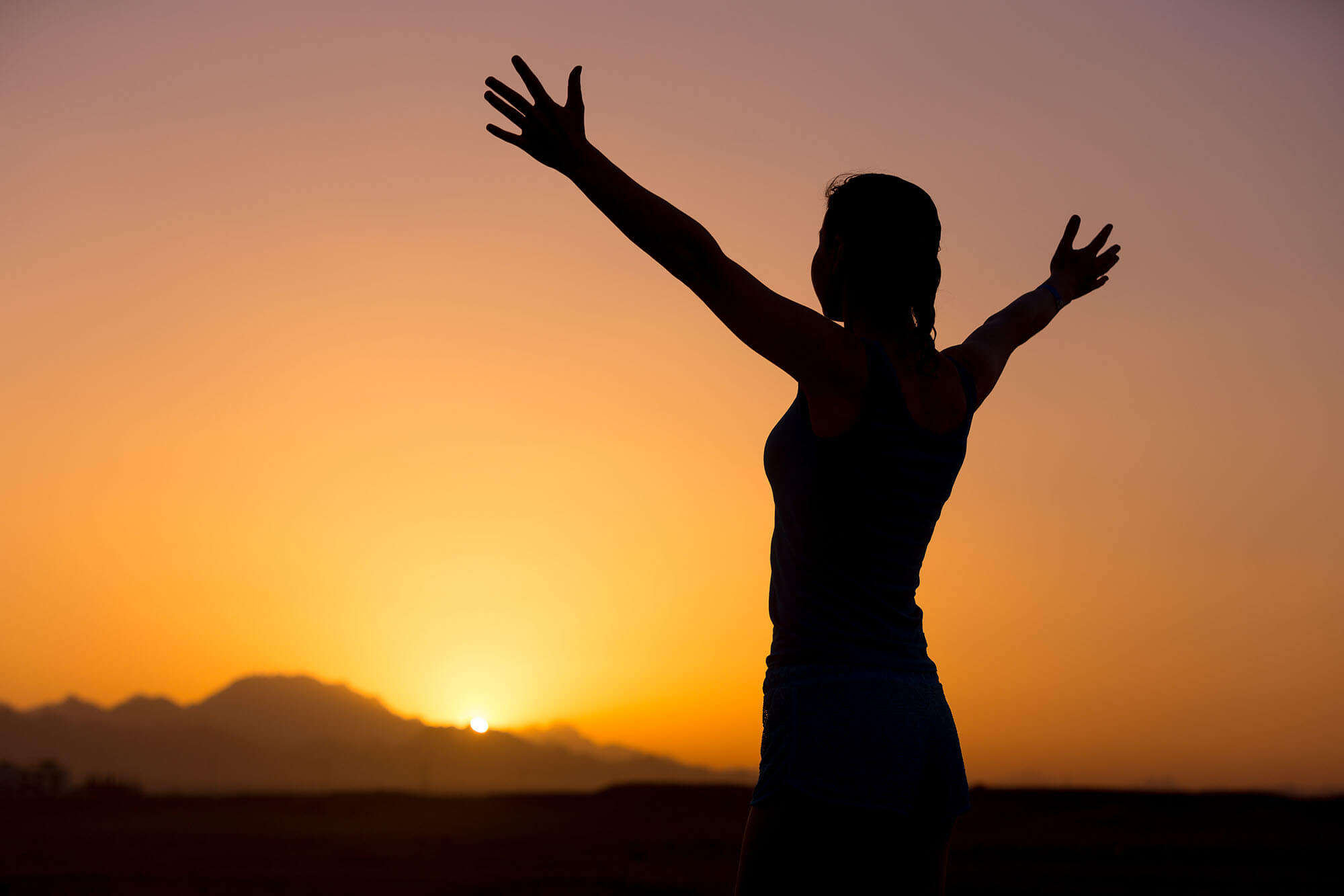A woman in silhouette with outstretched arms looking at the sunset.