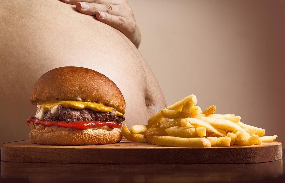 An image of a man's bare bulging stomach beside a table with a hamburger sandwich and French fries.