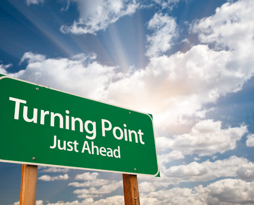 An image of a large green street sign under a blue sky with text that reads "turning point just ahead."