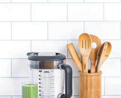 An image of a pitcher, a contain of wooden cooking and serving spoons, and a glass of green smoothie in front of kitchen wall tiles.