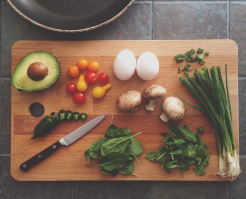 An image of a cutting board with green onions, whole eggs, grape tomatoes, mushrooms, baby spinach leaves, and a avocado half.
