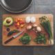 An image of a cutting board with green onions, whole eggs, grape tomatoes, mushrooms, baby spinach leaves, and a avocado half.