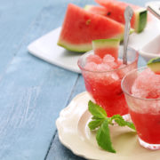 An image of watermelon wedges beside two glass of watermelon beverages on a table.