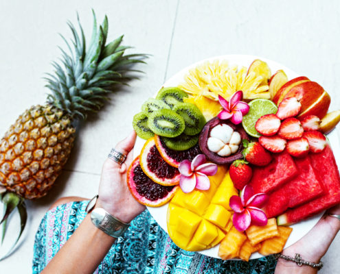 An image of hands holding a place of sliced kiwi, pineapple, strawberries, apple, pomegranate and mangosteen fruit.