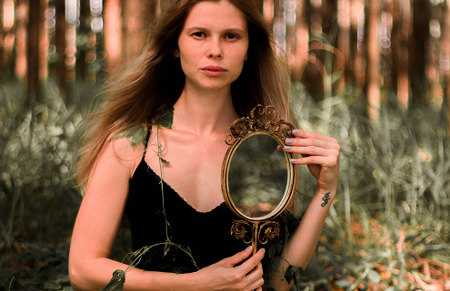 An image of an attractive woman in the forest holding a hand mirror.