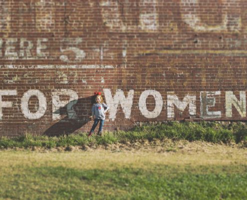An image of a little girl standing in front of a wall with white letters that spell "for women."