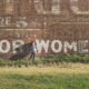 An image of a little girl standing in front of a wall with white letters that spell "for women."