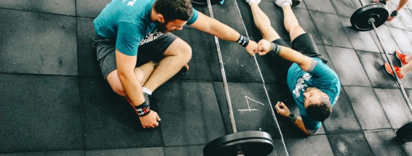 An image of two men sitting on the floor of a gym fist-bumping over a barbell.