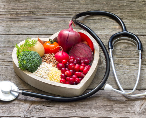 An image of a heart-shaped bowel full of healthy foods, including broccoli, onions, and tomatoes, beside a stethoscope on a table.
