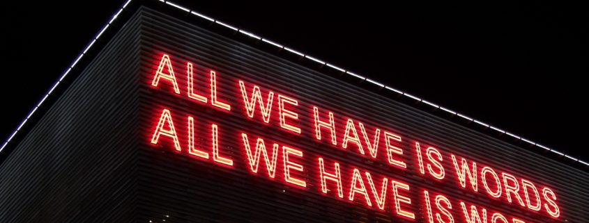 A neon sign on a building that reads "All we have is words, All we have is world."