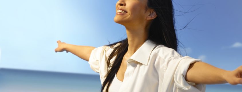 An image of a smiling woman on a beach looking toward the sky with outstretched arms.