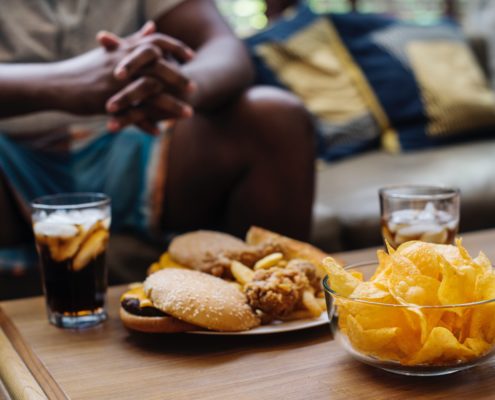 An image of a glass of soda, a plate of hamburgers, and a bowl of potato chips on table with a man sitting in the background.