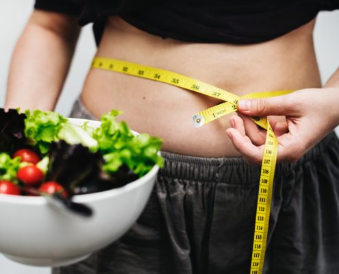 Image of a female with a measuring tape wrapped around her belly and a bowl of salad in hand. Prevent weight gain with the SANE Diet.
