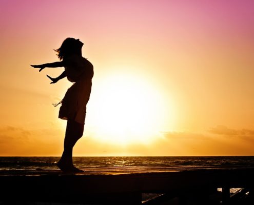 An image of a silhouette of a woman stretching her arms behind her while looking up at the sky at sunset.
