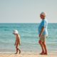 An image of an obese elderly man and his little grandson in matching hats walking on the beach.