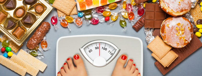 An image of female feet on a bathroom scale surrounded by junk foods, including candy, chocolate squares, donuts, and crackers.