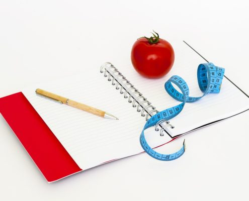 An image of an open notebook with ink pen, a tomato, and a measuring tape laying on top of it.