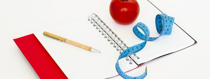 An image of an open notebook with ink pen, a tomato, and a measuring tape laying on top of it.