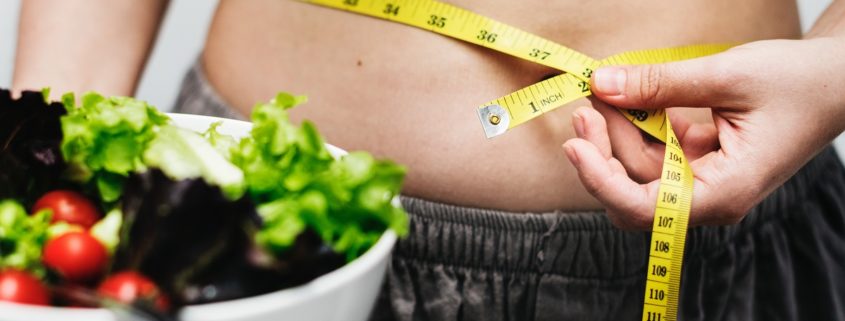 An image of a woman, one hand holding a tape measure around her waist, the other hand holding a salad.
