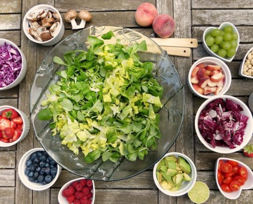 An image of a bowl of salad greens surrounded with bowls of chopped fruits and vegetables to add.