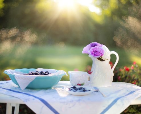 An image of a bowl of blueberries and a pitcher of flowers sitting on a small table outside. 