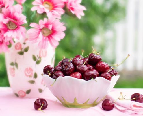 An image of pink flowers in a vase beside a bowl of cherries on a table with pink tablecloth.