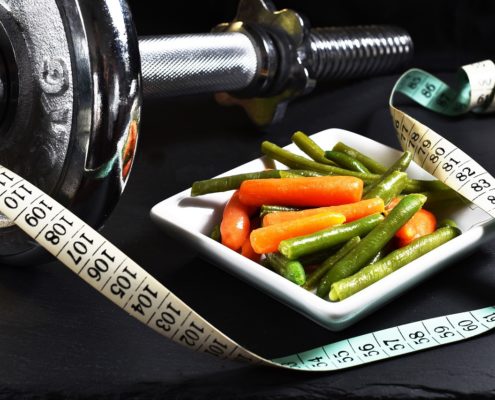 An image of a bowl of steamed green beans and carrots beside a tape measure and barbell.