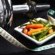 An image of a bowl of steamed green beans and carrots beside a tape measure and barbell.