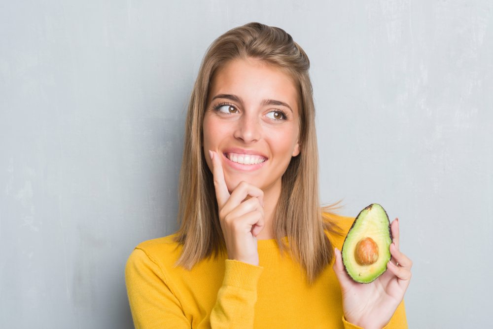 An image of a happy young woman holding an avocado half with pit to the camera.