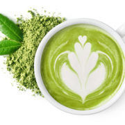 An image of a mug of green tea, with green tea powder and leaves beside it on white background.
