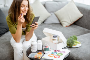 An image of a happy young woman on a sofa with a plate of broccoli and supplement pill bottles on the table in front of her. 