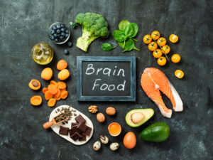 An image of salmon, avocado, dark chocolate, blueberries, and other foods with a small chalkboard that reads: brain foods. 