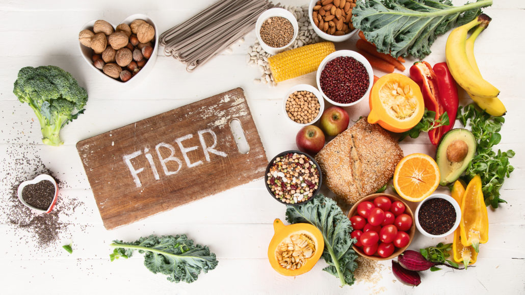 An image of high-fiber foods, such as walnuts, squash, and broccoli, surrounding a wooden sign that reads, "fiber."