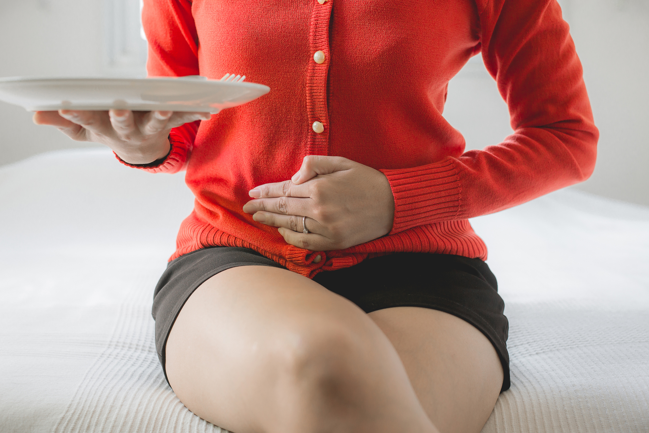 An image of a woman sitting on a bed holding her stomach in pain after eating.