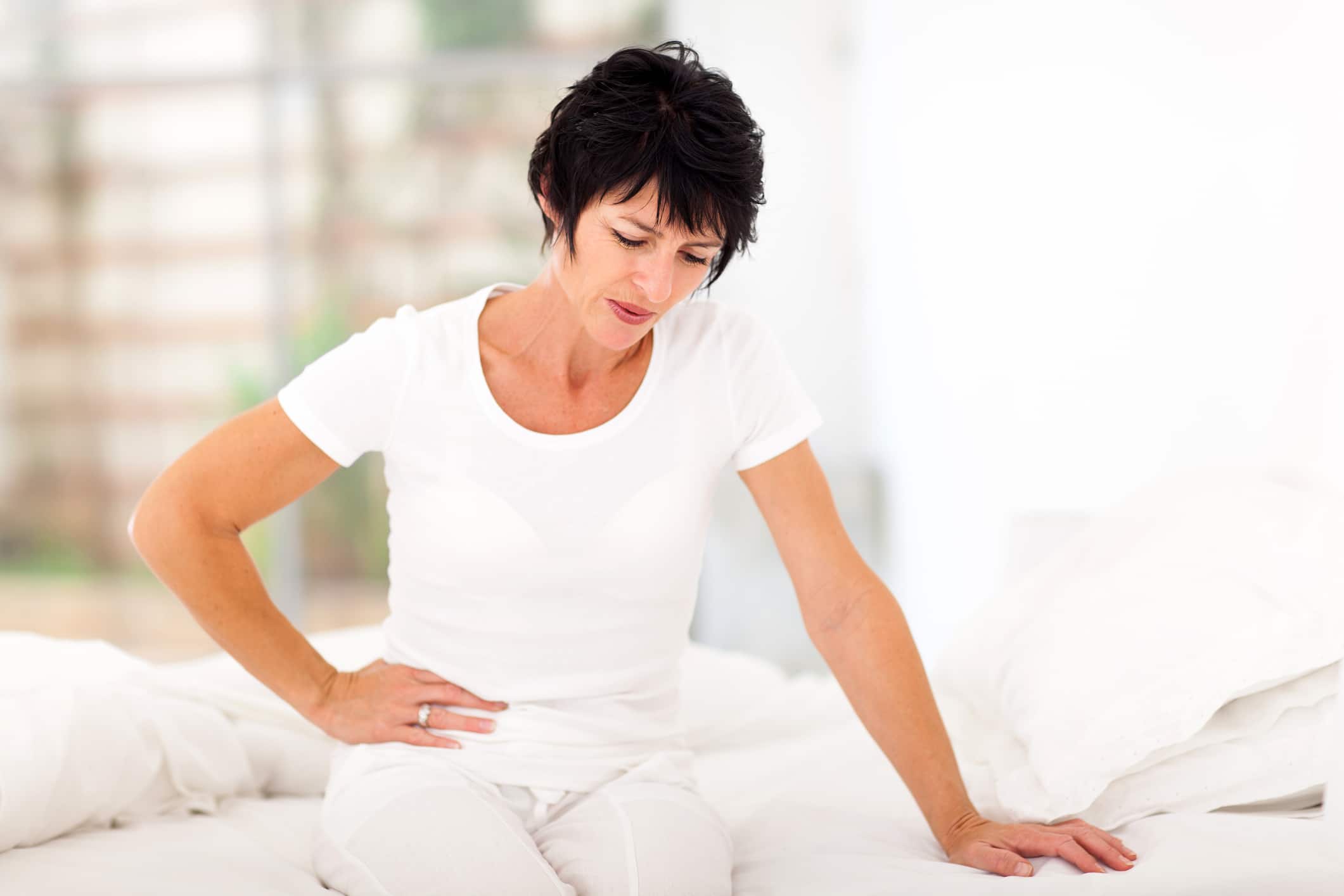 An image of a mature woman with a stomach ache sitting on a bed.