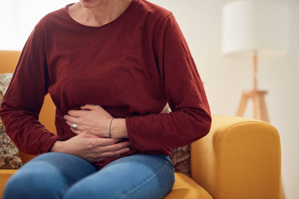 An image of a woman sitting on a sofa and holding her stomach in pain.