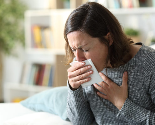 An image of an adult woman coughing into a tissue at home.