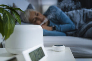 An image of a humidifier emitting steam on nightstand while a woman sleeps in bed.