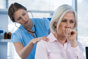 An image of mature woman coughing while doctor listens to her lungs in her back.